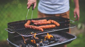 Person wendet Würstchen auf einem Grill, während Flammen darunter auflodern.